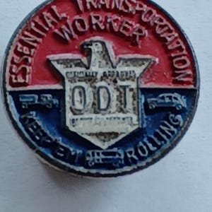 May include: A round pin with a red and blue border, featuring the text "ESSENTIAL TRANSPORTATION WORKER" and "KEEP 'EM ROLLING." The center has a white shield with "ODT" and an eagle.