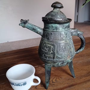 May include: A green bronze teapot with intricate carvings and a dragon-shaped spout. The teapot has three legs and a lid with a knob on top. It is sitting on a wooden surface next to a white cup with a blue floral design.