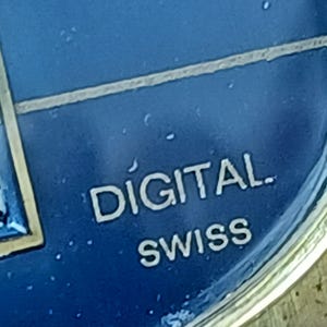Puede incluir: Primer plano de una esfera de reloj con un fondo azul oscuro. La palabra "DIGITAL" est&aacute; impresa en blanco, encima de "SWISS". La esfera del reloj tiene un borde met&aacute;lico.