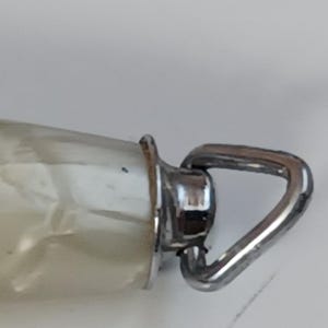 May include: Close-up of a clear glass bottle with a silver metal loop. The bottle has a pearlescent, off-white interior. The metal loop is attached to a silver metal collar.