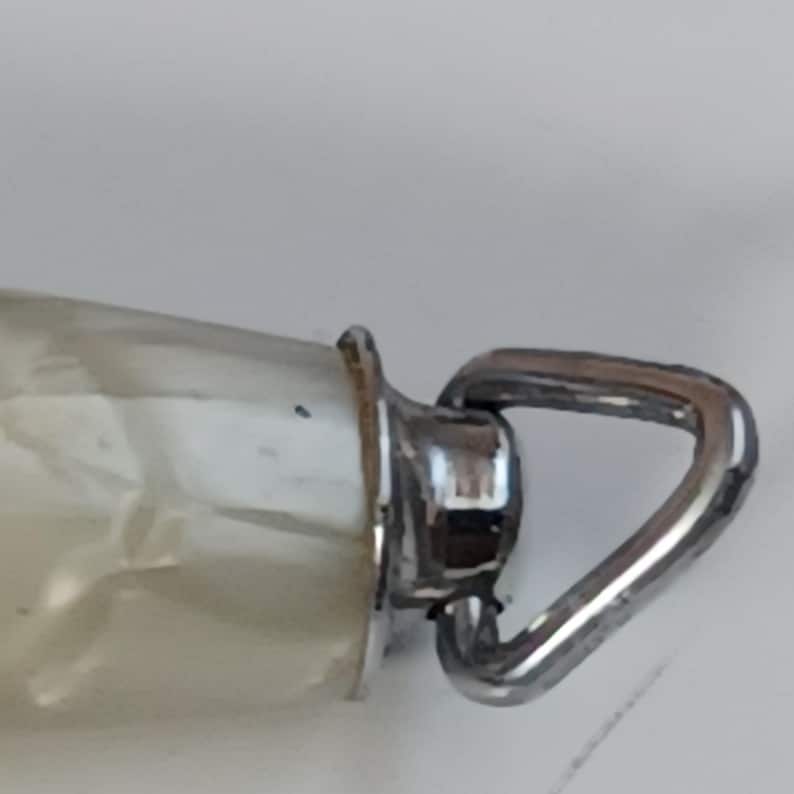 May include: Close-up of a clear glass bottle with a silver metal loop. The bottle has a pearlescent, off-white interior. The metal loop is attached to a silver metal collar.