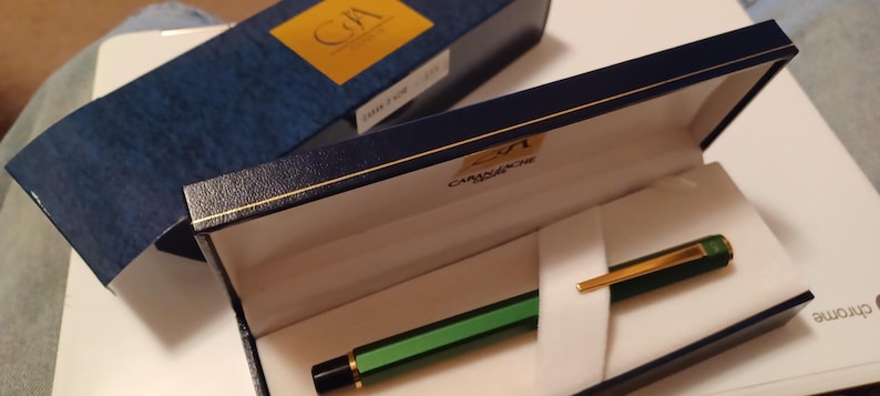 Puede incluir: Una pluma estilogr&aacute;fica verde y dorada en una caja azul con el texto "2/1 CAMISADO" en el interior de la tapa de la caja.