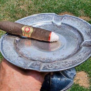 May include: A silver ashtray with an ornate design and a brown cigar resting on top. The cigar has a red and white band with the text "Romeo y Julieta" on it.