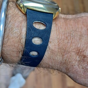 Puede incluir: Primer plano de un reloj de pulsera dorado con una correa de cuero azul oscuro. La correa presenta tres cortes ovalados. Una pulsera plateada es visible en la mu&ntilde;eca.