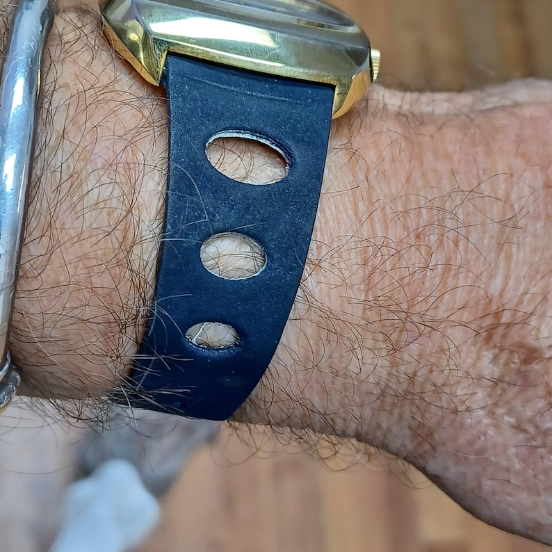 Puede incluir: Primer plano de un reloj de pulsera dorado con una correa de cuero azul oscuro. La correa presenta tres cortes ovalados. Una pulsera plateada es visible en la mu&ntilde;eca.