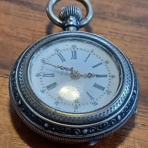 May include: A silver pocket watch with a white face and black Roman numerals. The watch has a decorative bezel and a winding stem with a loop. The hands are black and the watch is set against a wooden surface.