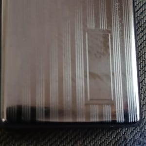 May include: A rectangular silver-toned metal case featuring a polished finish with a series of vertical engraved lines and a central rectangular frame detail. The surface is reflective and smooth, showcasing a clean, metallic aesthetic throughout.