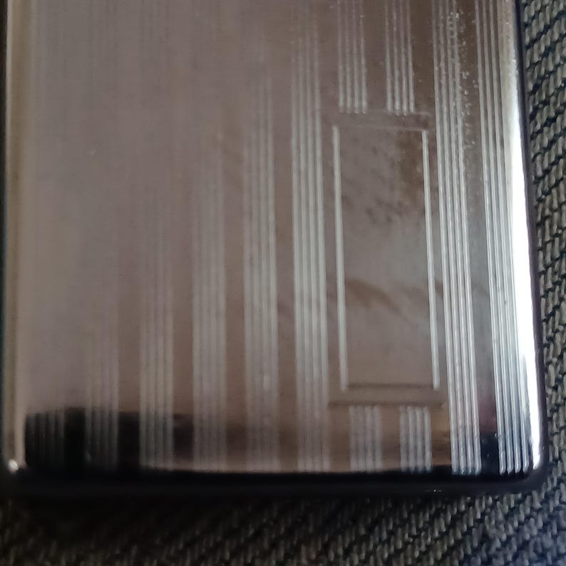 May include: A rectangular silver-toned metal case featuring a polished finish with a series of vertical engraved lines and a central rectangular frame detail. The surface is reflective and smooth, showcasing a clean, metallic aesthetic throughout.