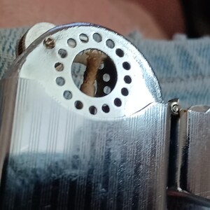 May include: A silver-toned metal lighter featuring a polished finish with vertical pinstripe detailing. The top section displays a circular wind guard with a series of small perforations surrounding a central wick, showing a metallic construction.