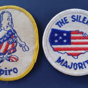 May include: Two embroidered patches. The first patch is yellow with a white background and features a cartoon figure wearing a red, white, and blue outfit. The figure is holding up two fingers and the text "Spiro" is below the figure. The second patch is white with a blue outline and features a blue and red American flag in the shape of the United States. The text "The Silent Majority" is around the flag.