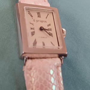 May include: A vintage silver-toned wristwatch with a rectangular face and a light pink leather band. The watch face displays Roman numerals and the text "à propos". The watch has a small crown and a textured band.
