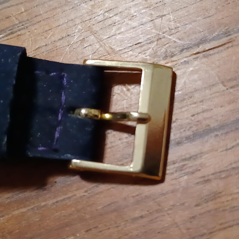 Puede incluir: Primer plano de una hebilla de cintur&oacute;n rectangular dorada con un pasador dorado. La hebilla est&aacute; sujeta a un cintur&oacute;n de cuero azul oscuro. El fondo es una superficie de madera.