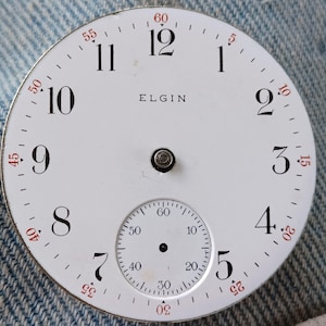 May include: A round white watch dial with black Arabic numerals from 1 to 12 and red minute markers in increments of five. The word ELGIN is printed in black in the center. A small sub-dial for seconds is positioned at the bottom with black markings.