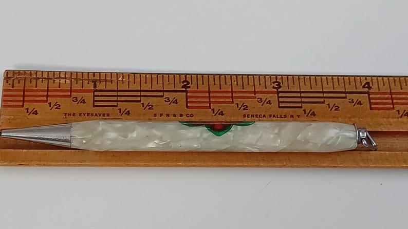 May include: A vintage mechanical pencil with a pearlescent white barrel and silver accents. The pencil has a decorative design with red and green details. It is placed next to a wooden ruler.