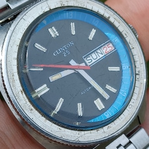 May include: A black and silver wristwatch with a blue bezel and a white face. The watch face has a date window that reads "SUN 25" and the text "CLINTON 25" and "AUTOMA".