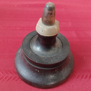 May include: A vintage bottle stopper with a dark brown base and a light brown top. The stopper has a cylindrical shape with a rounded top and a clear plastic ring. The base is circular and has a slightly textured surface.
