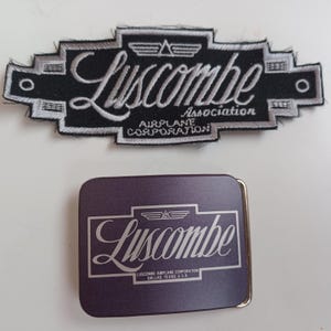 May include: A black and white embroidered patch with the text "Luscombe Airplane Association Corporation" and a silver belt buckle with the text "Luscombe Airplane Corporation Dallas, Texas U.S.A."