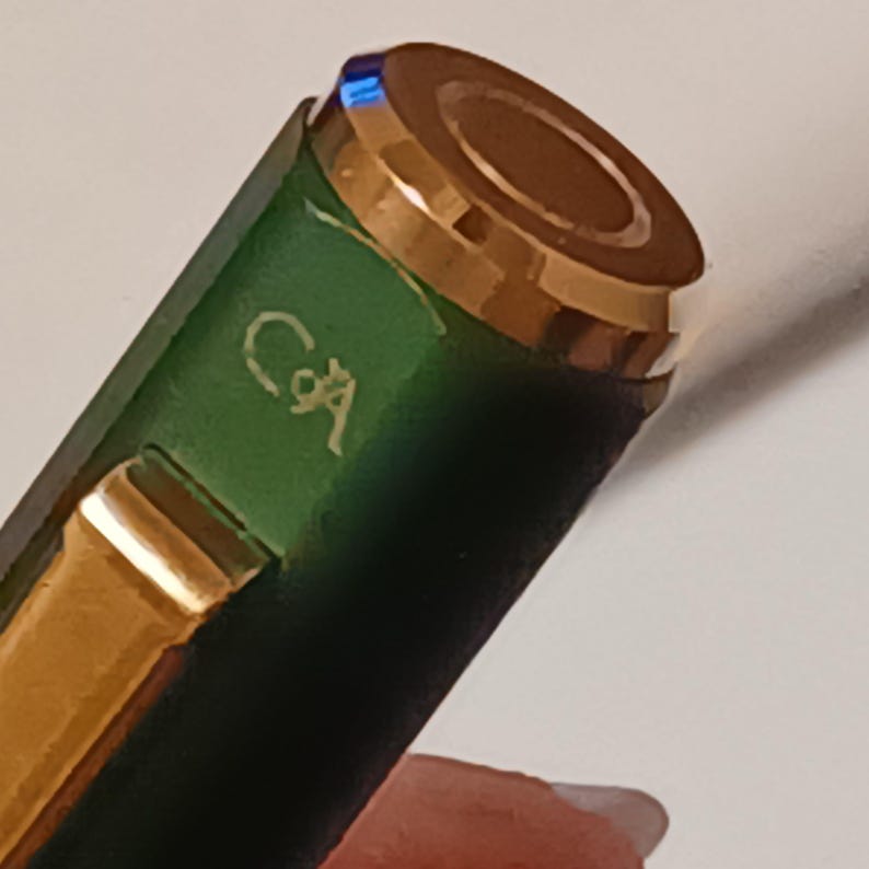 Puede incluir: Primer plano de una pluma verde con detalles dorados. La pluma tiene un capuch&oacute;n dorado con la parte superior marr&oacute;n y las letras "C&A" grabadas en el lateral.