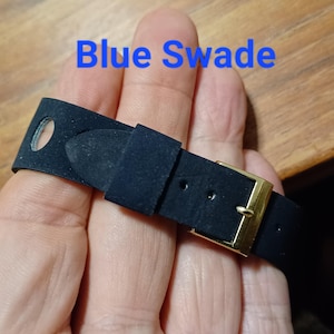 Puede incluir: Una correa de reloj de ante azul oscuro con una hebilla dorada. El texto "Blue Swade" est&aacute; escrito en azul en la parte superior de la imagen. La correa est&aacute; sujeta por una mano.