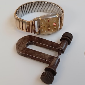 May include: A gold-toned wristwatch with a square face and a red accent on the dial. The watch is on a gold-toned metal band. A rusty metal tool is also in the image.