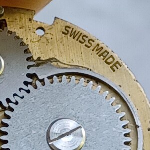 May include: Close-up of a vintage watch movement. The image shows a gold-toned metal plate with the words "SWISS MADE" engraved on it. A silver-colored gear with intricate teeth is also visible, along with a small screw.
