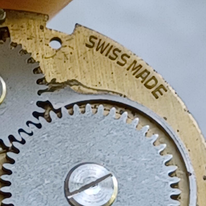 May include: Close-up of a vintage watch movement. The image shows a gold-toned metal plate with the words "SWISS MADE" engraved on it. A silver-colored gear with intricate teeth is also visible, along with a small screw.