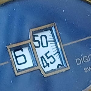 Puede incluir: Primer plano de una esfera de reloj con una pantalla digital que muestra la hora. La pantalla muestra el n&uacute;mero 6 y los n&uacute;meros 50 y 45. El fondo es de color azul oscuro.