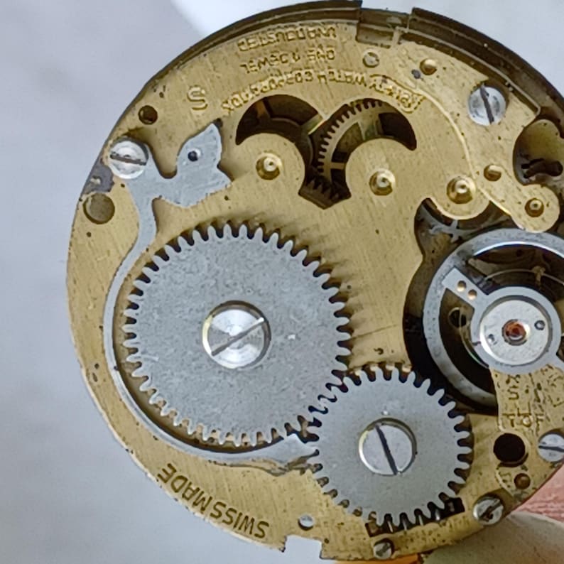 May include: Close-up of a watch movement with visible gears and intricate mechanical components. The watch mechanism features a silver-colored balance wheel and two large, silver-colored gears. The words "Swiss Made" are visible.