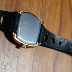 Puede incluir: Un reloj dorado con una esfera rectangular y una correa de cuero negro. El reloj tiene una parte trasera plateada y una peque&ntilde;a corona. La correa tiene varios agujeros para ajustar.