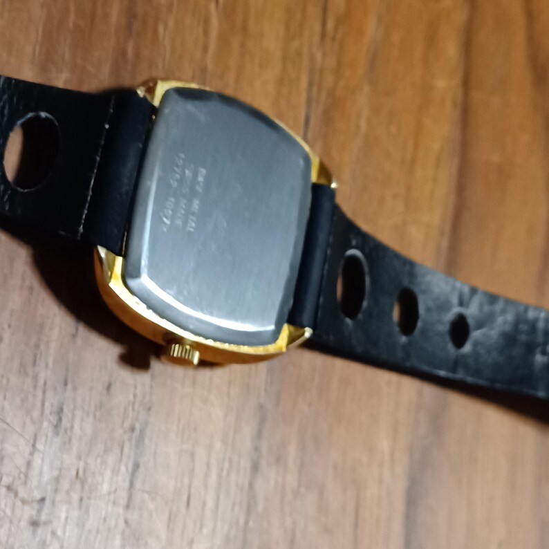 Puede incluir: Un reloj dorado con una esfera rectangular y una correa de cuero negro. El reloj tiene una parte trasera plateada y una peque&ntilde;a corona. La correa tiene varios agujeros para ajustar.