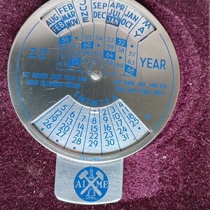 May include: A silver-colored metal calendar wheel with a blue and white design. The wheel has a rotating dial with the months of the year and days of the week. The wheel also has a small dial for setting the year. The text "SET MONTH OVER YEAR AND READ CALENDAR BELOW" and "USE DARK JAN. AND FEB. FOR LEAP YEARS ONLY" is printed on the wheel. The wheel has a small metal tag with the text "AIME" and a crossed pickaxe design.