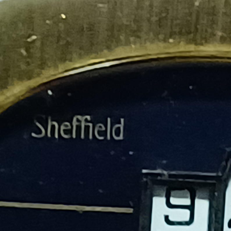 Puede incluir: Primer plano de una esfera de reloj con el nombre "Sheffield" en letras plateadas. La esfera es azul oscuro con un bisel dorado. El n&uacute;mero "9" es visible en blanco.