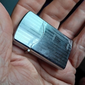 May include: A rectangular silver-tone metal lighter featuring a polished finish with a series of fine, horizontal engraved lines across the surface. The lighter has a hinged lid and a smooth, rounded top edge, reflecting light across its metallic body.