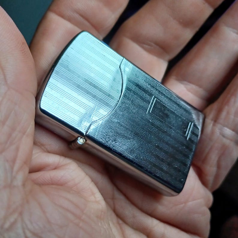 May include: A rectangular silver-tone metal lighter featuring a polished finish with a series of fine, horizontal engraved lines across the surface. The lighter has a hinged lid and a smooth, rounded top edge, reflecting light across its metallic body.