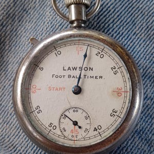 May include: A vintage Lawson Foot Ball Timer stopwatch with a white face, black numerals, and red accents. The stopwatch has a metal casing and a small subdial. The text "LAWSON FOOT BALL TIMER" is printed on the face.