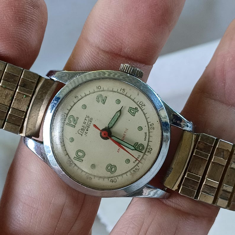 May include: A vintage Liberty Jeweled wristwatch with a silver-tone case and a cream-colored face. The watch features black numerals, green hands, and a red second hand. The watch has a gold-tone, expandable band.