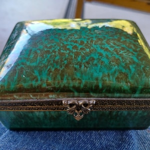 May include: A green ceramic trinket box with a brass hinged lid and decorative floral design. The box has a glossy finish.