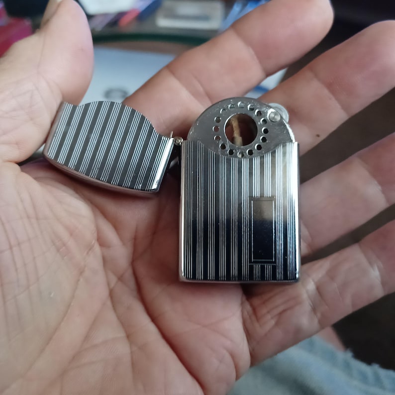 May include: A silver-toned metal lighter with a hinged lid, featuring a vertical pinstripe pattern across the body and lid. The top section of the lighter has a circular wind guard with small perforations and a visible wick in the center.