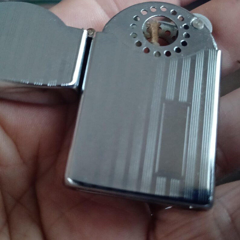 May include: A silver-toned metal lighter with a hinged lid and a rectangular body. The surface features a series of vertical etched lines and a central rectangular panel. The top section includes a circular wind guard with small perforations.