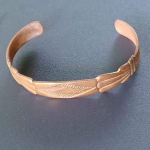 May include: A copper-colored cuff bracelet with an open design. The bracelet features a central diamond-shaped pattern with etched details and stylized feather-like designs on either side. The metal has a slightly aged appearance.