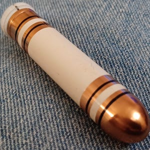 May include: A cylindrical object with a white body, accented with copper-colored bands and black stripes. The top and bottom are also copper-colored. The object is lying on a blue denim surface.