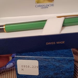 Puede incluir: Un bol&iacute;grafo Caran d'Ache verde y dorado en su caja azul. La caja tiene las palabras "Swiss Made" y el n&uacute;mero "0958.229 M" impresas en ella.