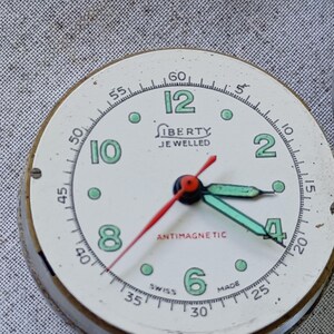 May include: Vintage Liberty watch face with a cream-colored dial, featuring black minute markers and green Arabic numerals. The watch has a red second hand and green hour and minute hands. The text "LIBERTY JEWELLED" and "ANTIMAGNETIC" are visible.