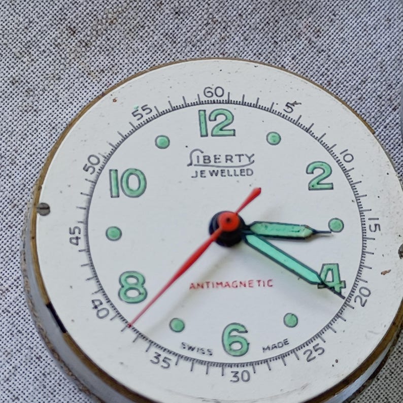 May include: Vintage Liberty watch face with a cream-colored dial, featuring black minute markers and green Arabic numerals. The watch has a red second hand and green hour and minute hands. The text "LIBERTY JEWELLED" and "ANTIMAGNETIC" are visible.