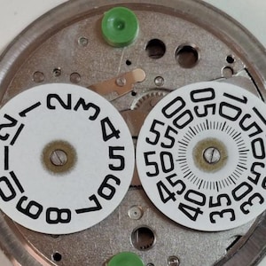 Puede incluir: Primer plano de componentes de reloj. Dos esferas circulares blancas con n&uacute;meros y marcas negras se colocan sobre una base de metal plateado. Tambi&eacute;n se ven piezas de pl&aacute;stico verde. Las esferas muestran n&uacute;meros para horas y minutos.