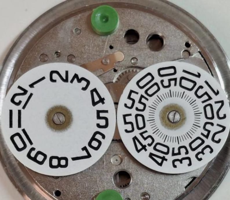 Puede incluir: Primer plano de componentes de reloj. Dos esferas circulares blancas con n&uacute;meros y marcas negras se colocan sobre una base de metal plateado. Tambi&eacute;n se ven piezas de pl&aacute;stico verde. Las esferas muestran n&uacute;meros para horas y minutos.