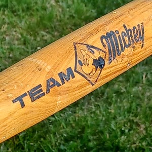 May include: A wooden baseball bat with the words "TEAM Mickey" printed on it. The bat has a black and white image of Mickey Mouse wearing a baseball cap.
