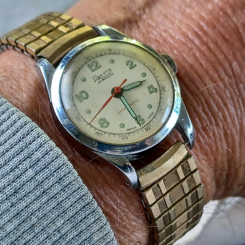 May include: Vintage Liberty 17 Jewels wristwatch with a cream-colored face, featuring black numerals and a red second hand. The watch has a silver-tone case and a gold-tone, expandable band. The watch is shown on a wrist.