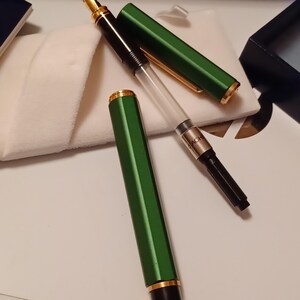 Puede incluir: Una pluma estilogr&aacute;fica verde con detalles dorados y un cartucho de tinta transparente. La pluma est&aacute; sobre una superficie blanca con un pa&ntilde;o blanco al lado.