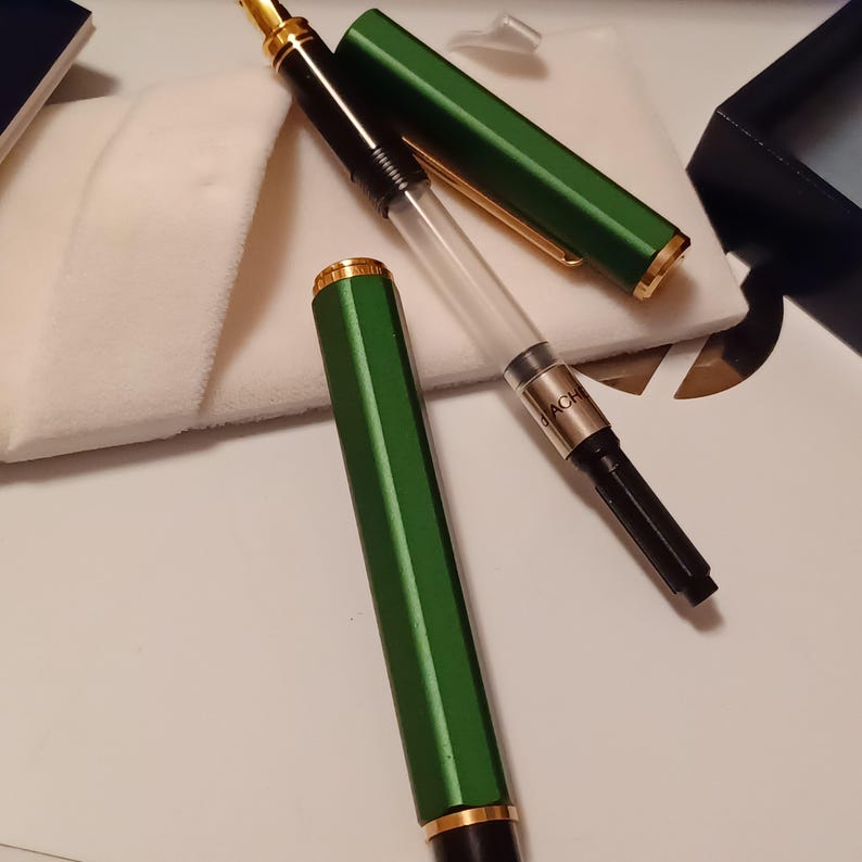 Puede incluir: Una pluma estilogr&aacute;fica verde con detalles dorados y un cartucho de tinta transparente. La pluma est&aacute; sobre una superficie blanca con un pa&ntilde;o blanco al lado.